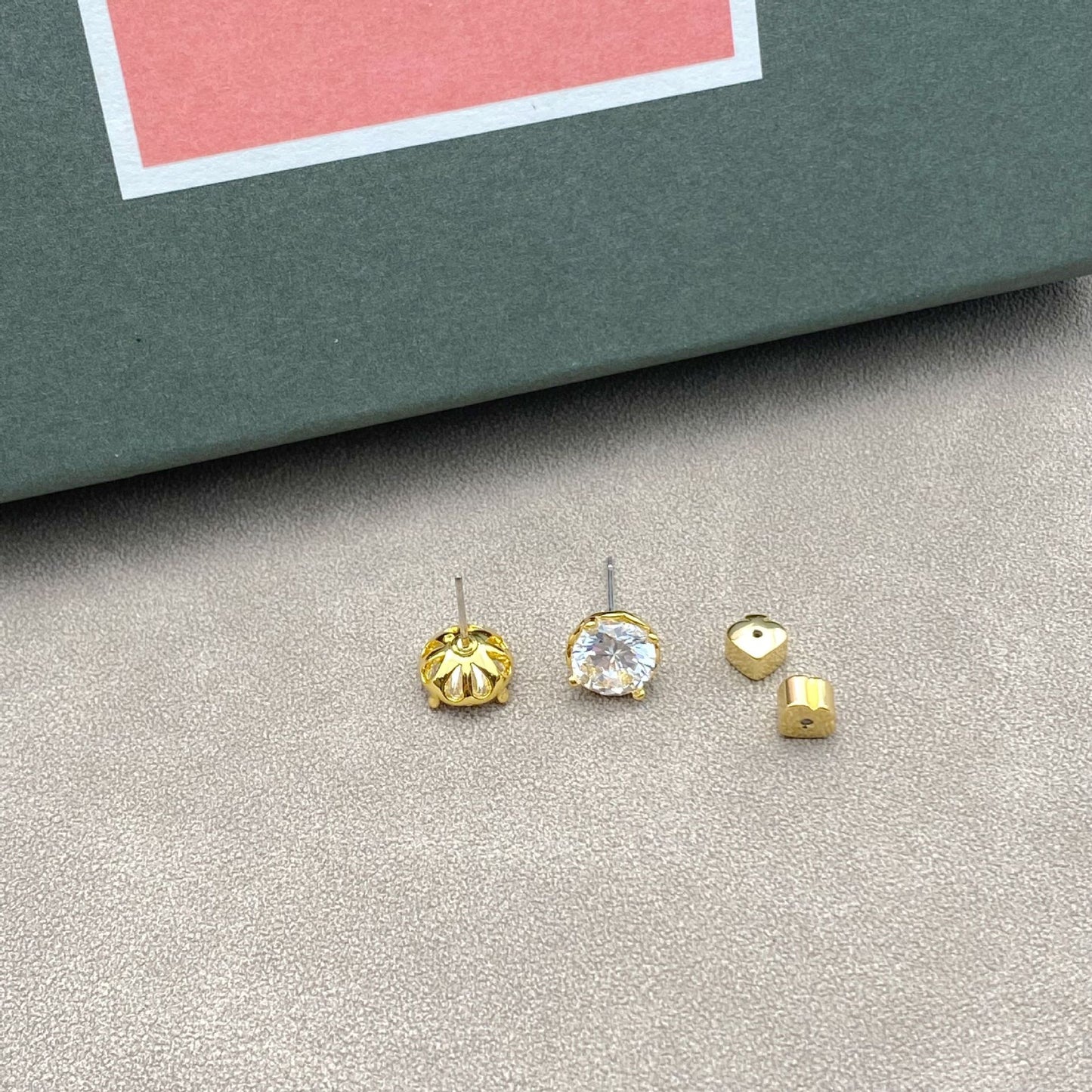 Gold and silver stud earrings on a gray surface with a pink and green box in the background.
