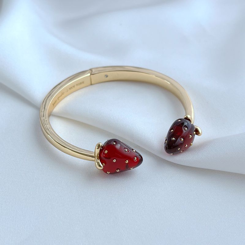 Bracelet on a light, natural background (white wood or pale pink), highlighting the strawberry enamel details. Alt: “Kate Spade Strawberry Gold Bracelet – Dainty Fruit Charm Jewelry.”