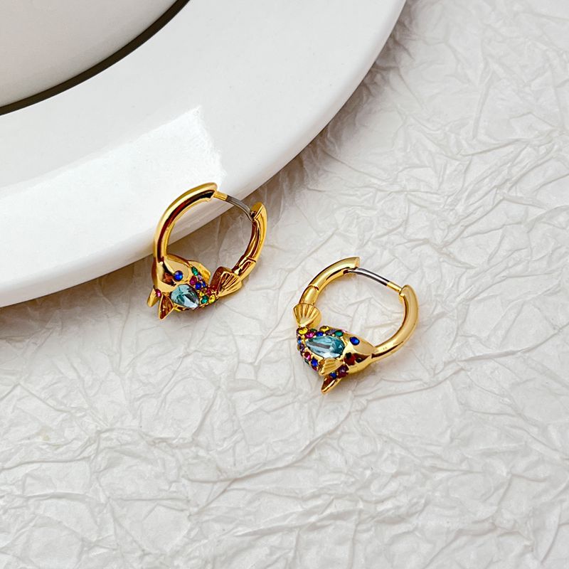 Earrings on a soft aquatic blue or seafoam green background, showcasing the gold huggie hoop with dangling enamel fish charm. Alt: “Kate Spade What A Catch Fish Huggie Earrings NEW – Gold Fish Hoop Jewelry.”
