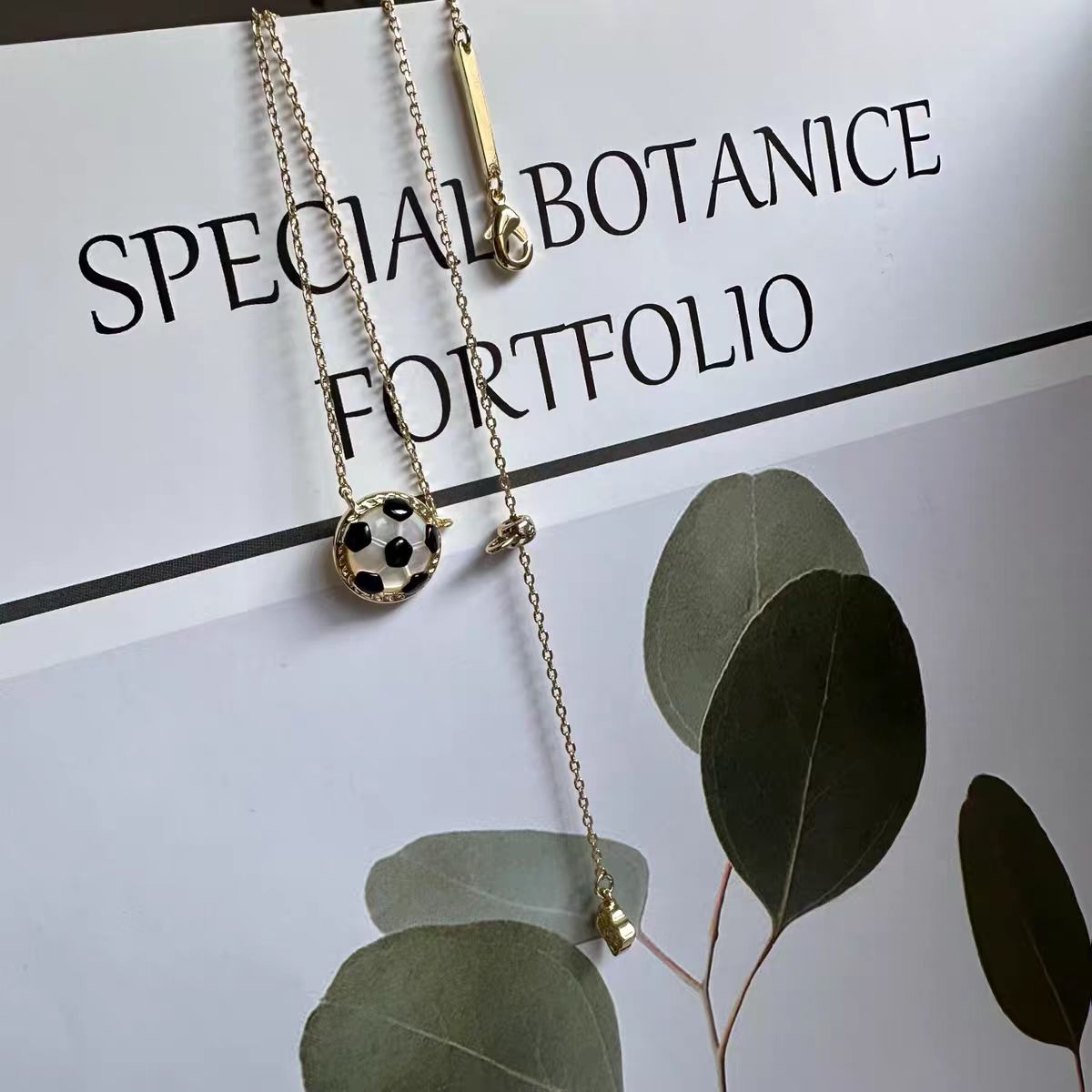 Necklace with a football pendant on a 'Special Botanice Portfolio' paper with green leaves.
