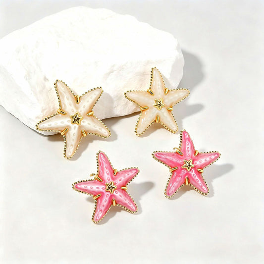 Four starfish-shaped stud earrings arranged on a white surface with a white rock prop. Two earrings are cream with gold trim and a small gold center, and two are pink with gold trim and a small gold center.