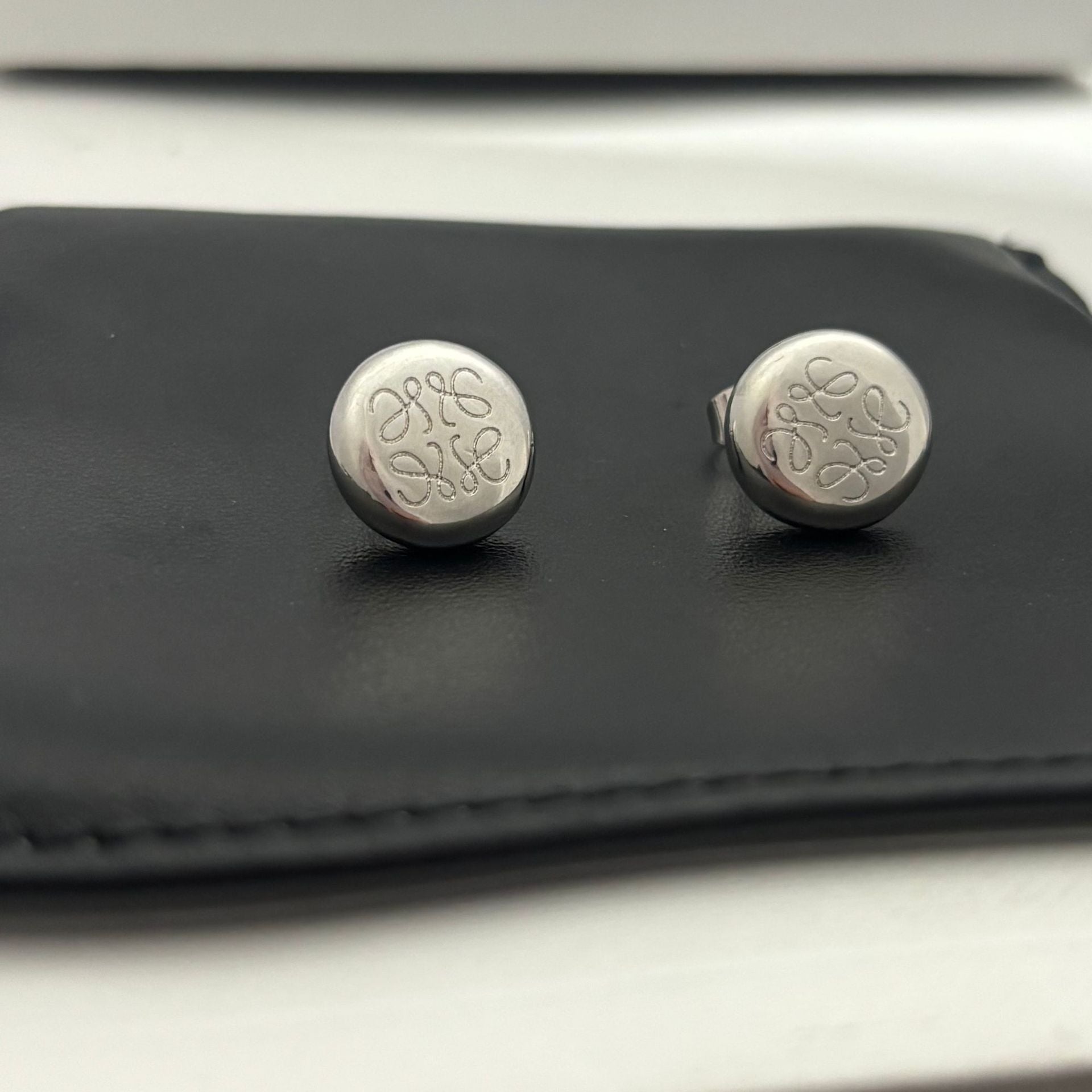 Clean shot of a single earring against a neutral, textured background (like linen or stone), highlighting the smooth pebble shape and integrated logo.