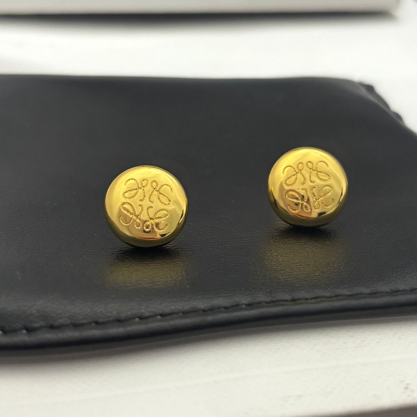 Clean shot of a single earring against a neutral, textured background (like linen or stone), highlighting the smooth pebble shape and integrated logo.