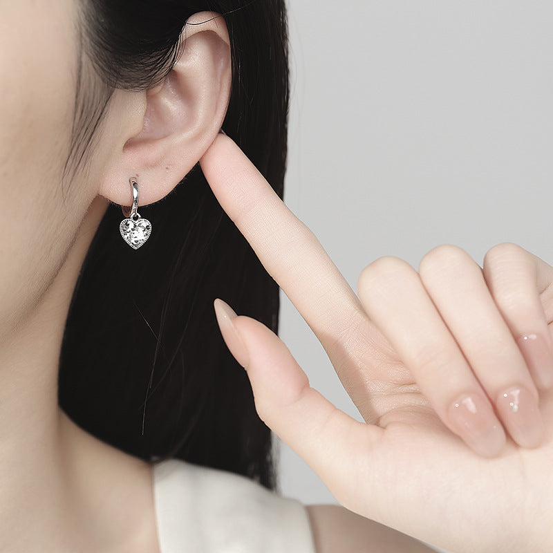 Heart-shaped earring being worn and held by a hand against a neutral background