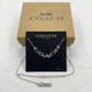 A silver-tone Coach letter necklace displayed on a black Coach card, with the necklace laid out in front and a tan Coach box behind it on a white background.