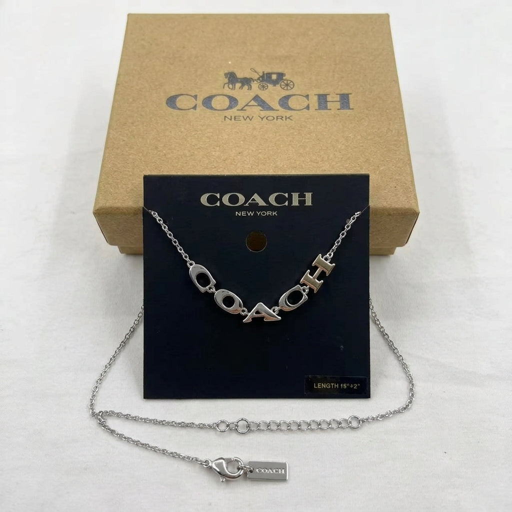 A silver-tone Coach letter necklace displayed on a black Coach card, with the necklace laid out in front and a tan Coach box behind it on a white background.