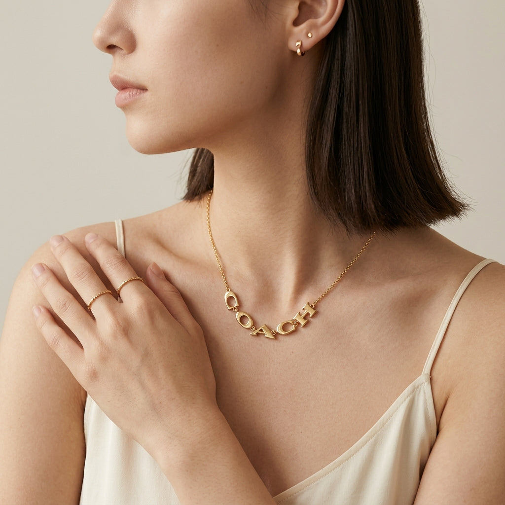 A close-up lifestyle shot of a woman wearing a gold initial necklace spelling 'COACH' across her chest, along with matching gold rings and earrings.