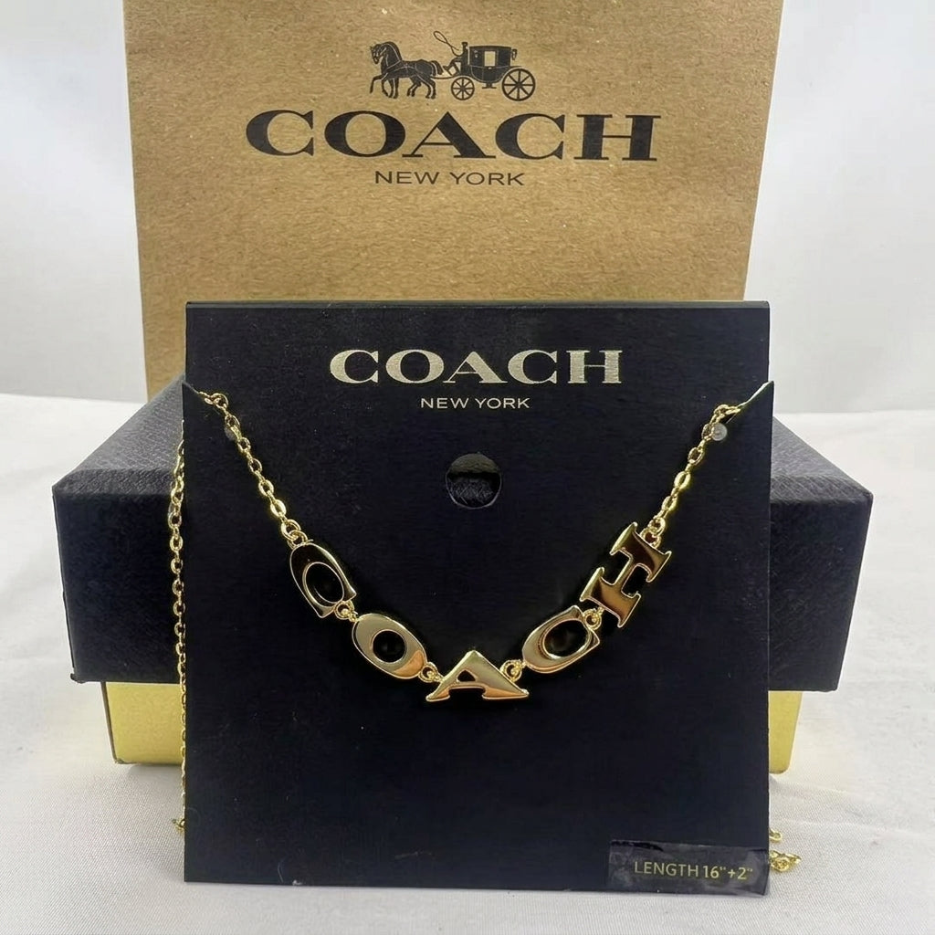 A gold-tone Coach logo chain necklace displayed on a black branded card, with a kraft Coach box behind it on a white background.