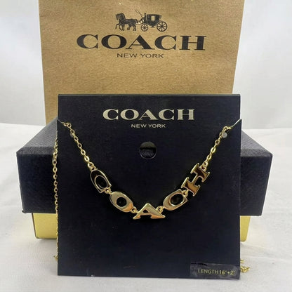 A gold-tone Coach logo chain necklace displayed on a black branded card, with a kraft Coach box behind it on a white background.