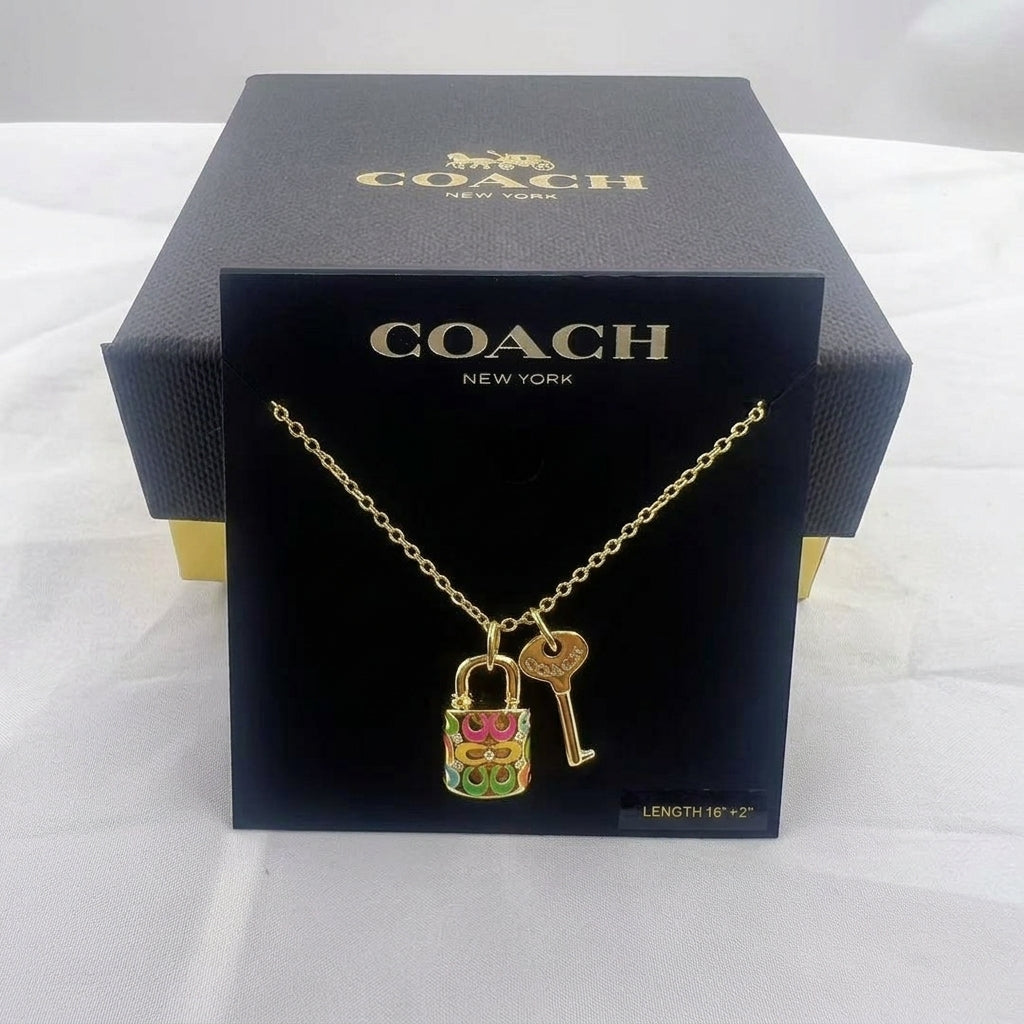 A gold-tone Coach necklace with a colorful enamel padlock and key pendant, displayed on a black Coach New York card in front of a black Coach box on a white fabric background.