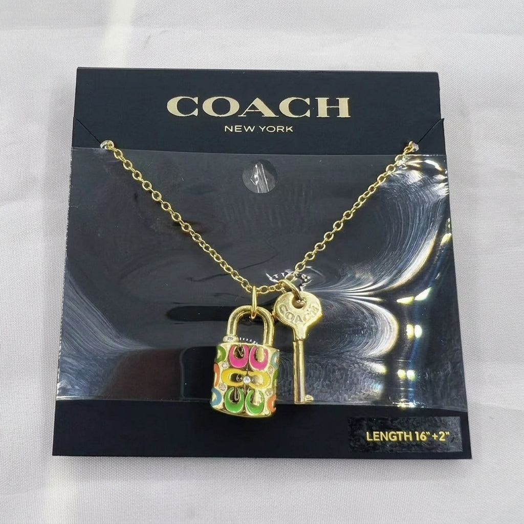 A gold-tone Coach necklace with a padlock and key pendant, displayed on its black branded backing card. The padlock features colorful Coach logo lettering in green, pink, and blue.