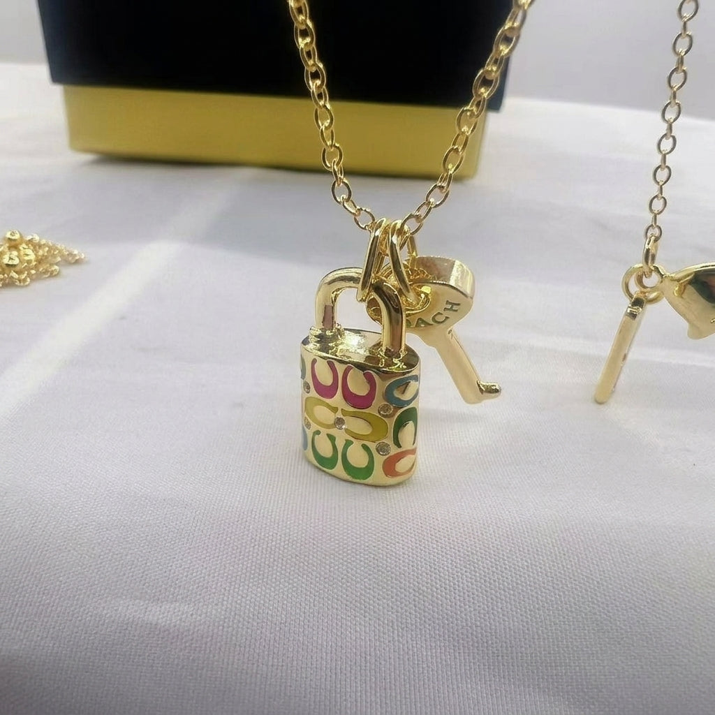 A gold-tone padlock pendant necklace with a colorful interlocking 'C' pattern, accompanied by a key charm, displayed on a white fabric surface.