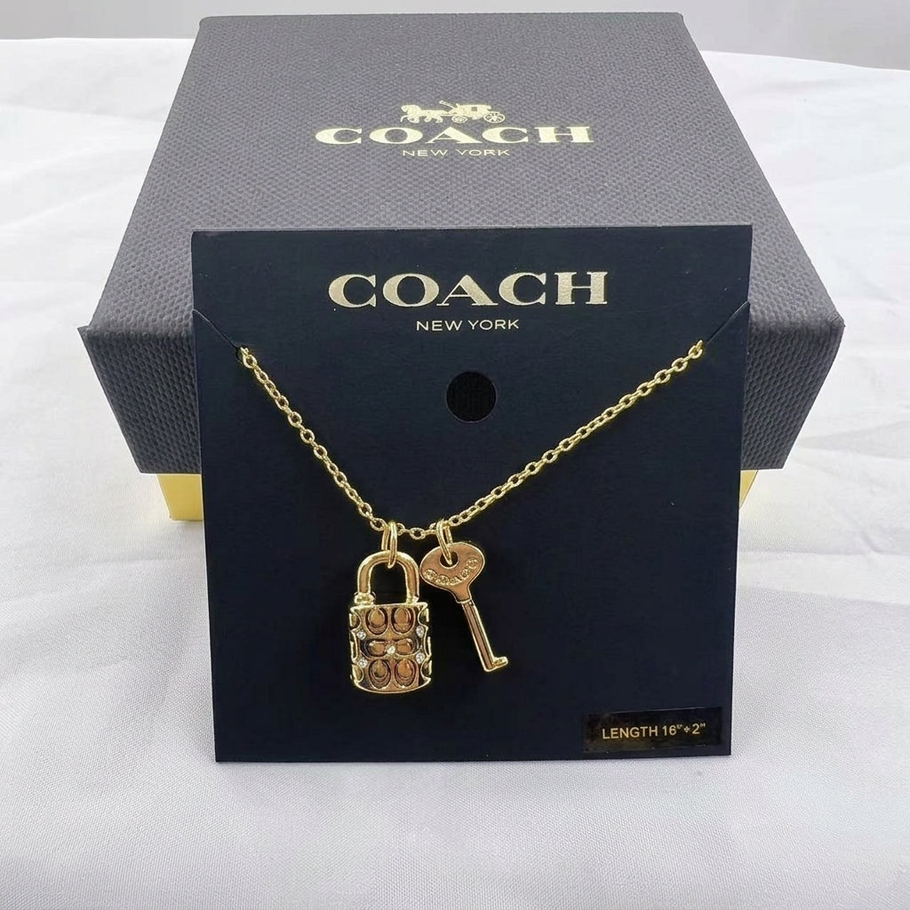 A gold-tone Coach necklace with a padlock and key pendant, displayed on a black Coach New York card in front of a dark gray Coach box on a white fabric background.