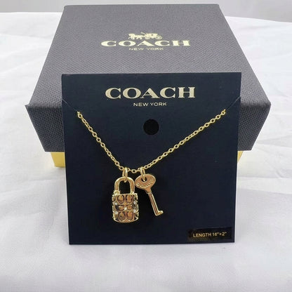 A gold-tone Coach necklace with a padlock and key pendant, displayed on a black Coach New York card in front of a dark gray Coach box on a white fabric background.