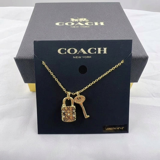 A gold-tone Coach necklace with a padlock and key pendant, displayed on a black Coach New York card in front of a dark gray Coach box on a white fabric background.