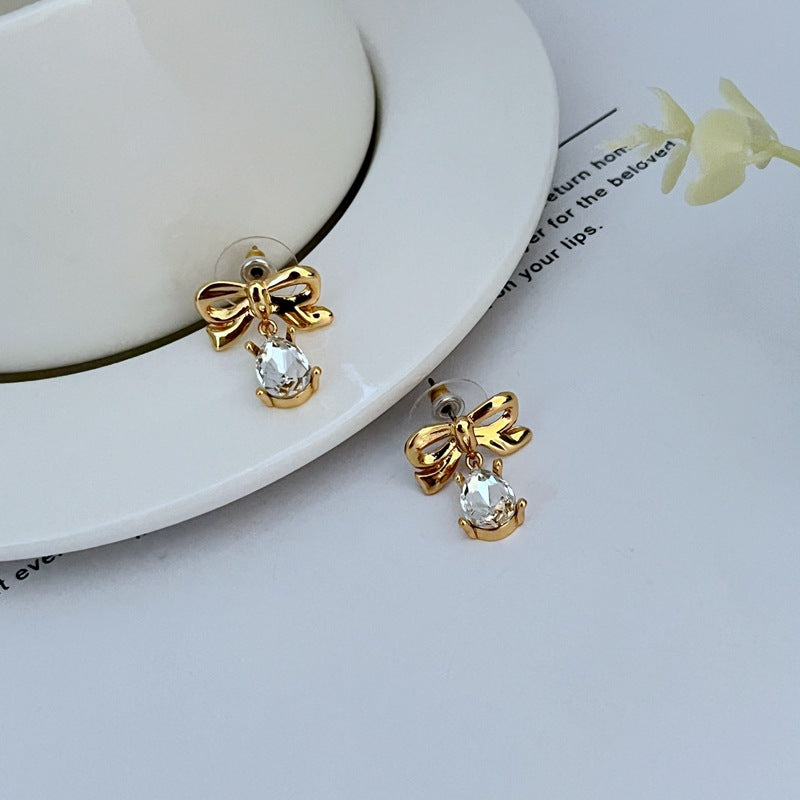 : Earrings on a soft blush pink or cream background, showcasing the teardrop silhouette and delicate bow detail. Alt: “New Kate Spade Gold Dainty Tied Up Tear Drop Earrings – Feminine Bow Knot Jewelry.”