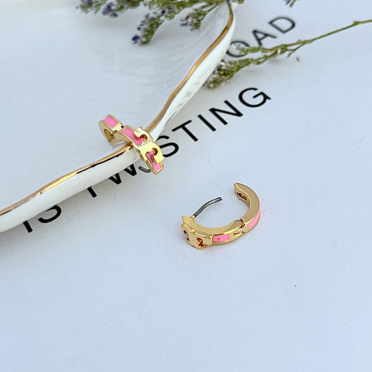 Both huggies displayed face-forward on a light background, highlighting the pink enamel T against the gold hoop.