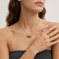 A close-up of a woman wearing a delicate gold chain necklace with a small cylindrical pendant and a matching gold ring on her finger, against a clean light background.