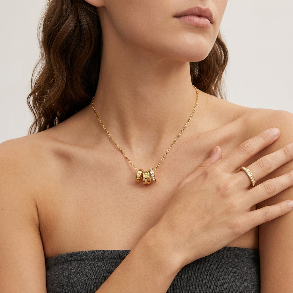 A close-up of a woman wearing a delicate gold chain necklace with a small cylindrical pendant and a matching gold ring on her finger, against a clean light background.