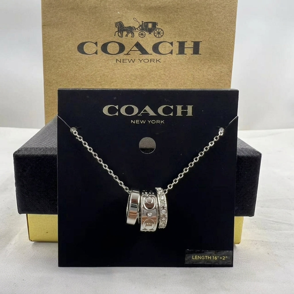 A Coach New York silver charm necklace displayed on a black branded card, with the gold-tone Coach box behind it on a white surface.