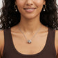 A close-up of a woman wearing a silver pendant necklace with three interlocking silver barbell-like charms, along with gold hoop earrings with white pearl drops, against a clean white background.
