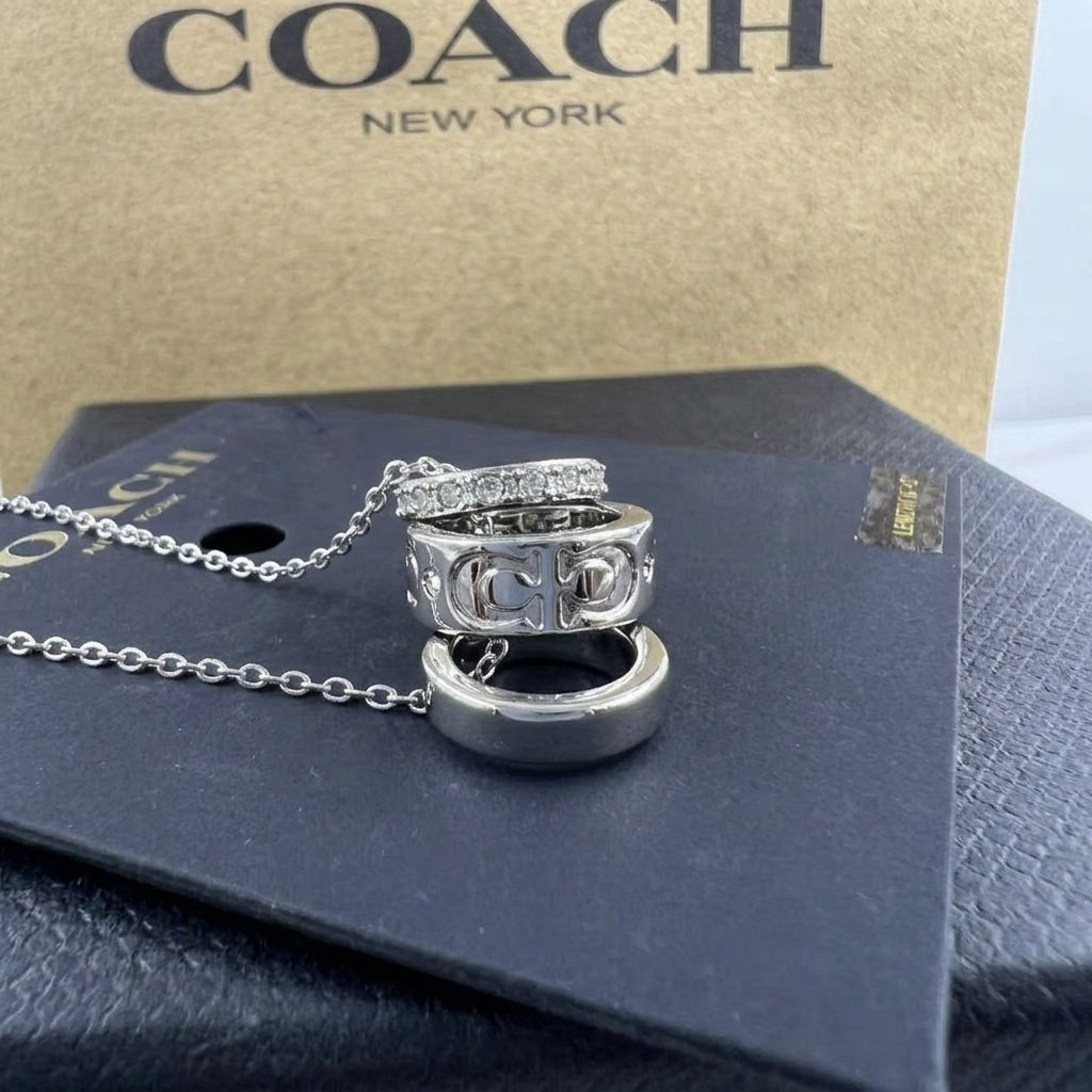 A Coach New York ring necklace with a silver-tone chain and three stacked rings featuring the interlocking CC logo, displayed on a black presentation card with a brown Coach box in the background.