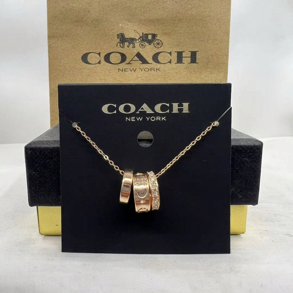 A Coach New York necklace with three interlocking gold-tone rings, one adorned with small clear crystals, displayed on a black branded card with a brown Coach box behind it.