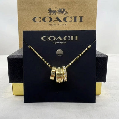 A gold-tone Coach necklace with three interlocking charm beads, displayed on a black COACH New York card in front of a brown COACH New York box.