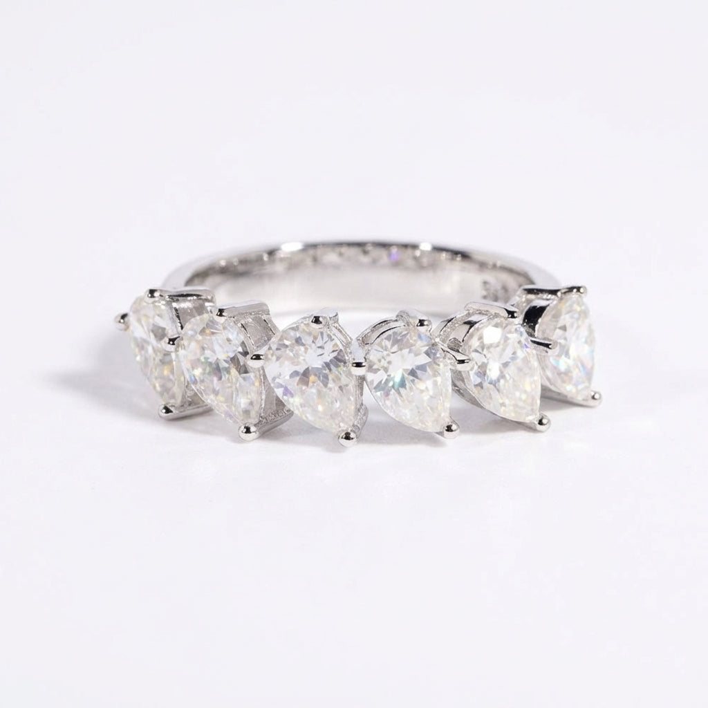 Clean white or soft pastel background — the pear shape slanted moissanite ring displayed on a flat surface or ring stand. The tilted pear-cut moissanite catches soft light from an angle, showcasing the unique asymmetrical orientation and brilliant sparkle.