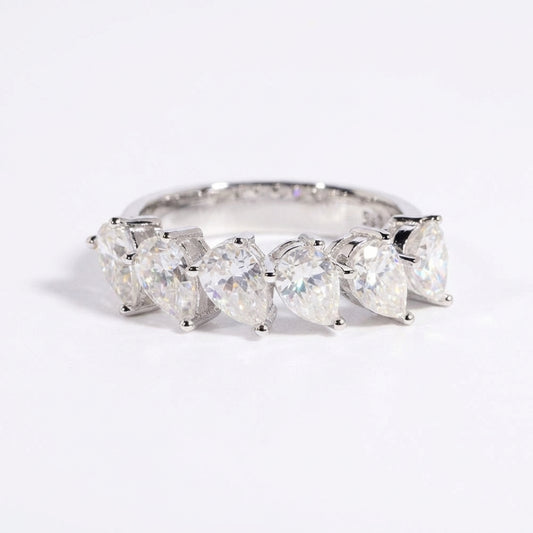 Clean white or soft pastel background — the pear shape slanted moissanite ring displayed on a flat surface or ring stand. The tilted pear-cut moissanite catches soft light from an angle, showcasing the unique asymmetrical orientation and brilliant sparkle.
