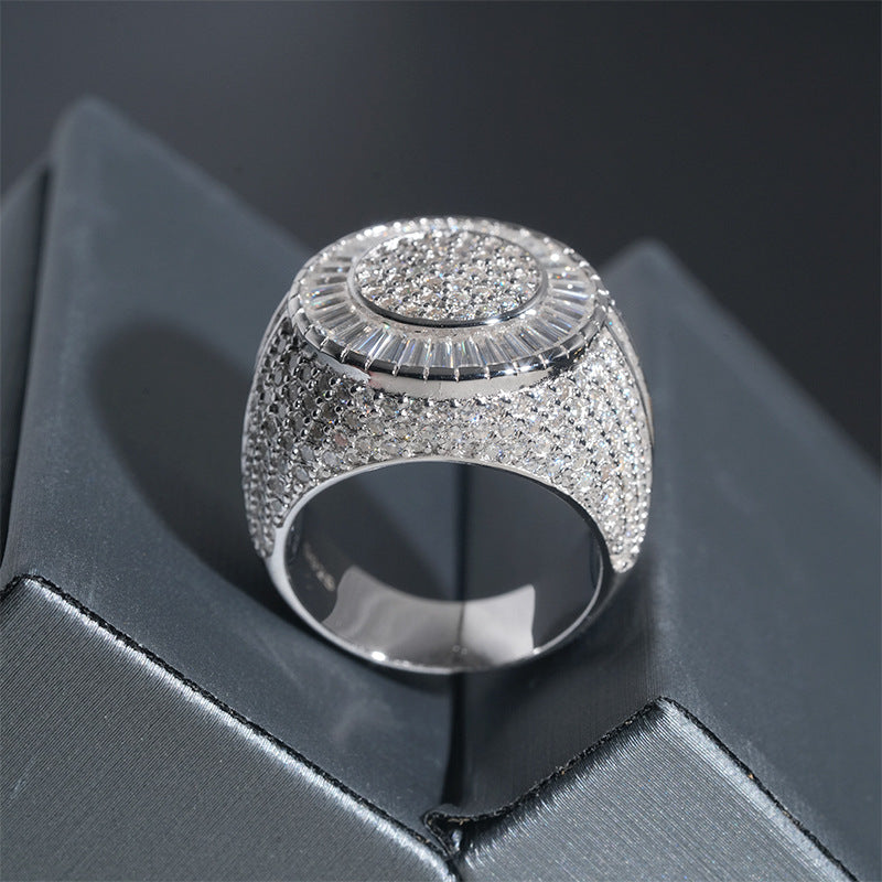 A silver-toned statement ring with a wide, heavily embellished pavé band and a circular textured top, displayed on a dark gray geometric surface.
