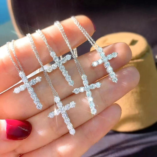 Four silver cross pendant necklaces, each set with clear crystals, held in a hand with red nail polish. A gold-toned jewelry box is blurred in the background.