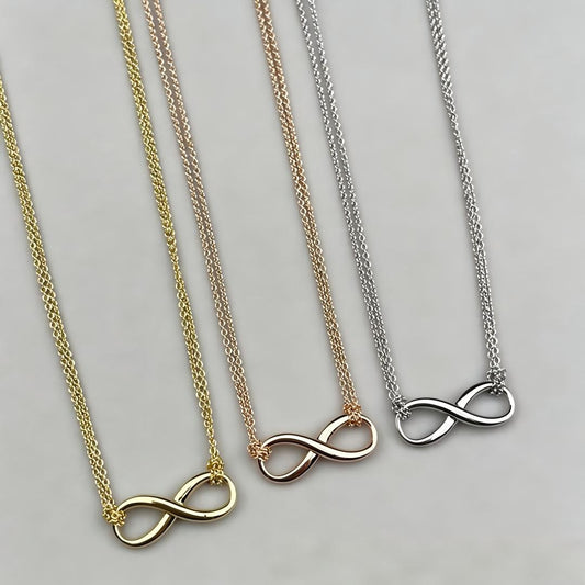 Three infinity necklace pendants on matching chains, shown in yellow gold, rose gold, and silver metal finishes on a light gray background.