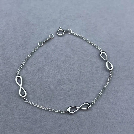 A silver chain bracelet featuring three polished infinity symbols spaced along the chain, displayed on a textured gray background.