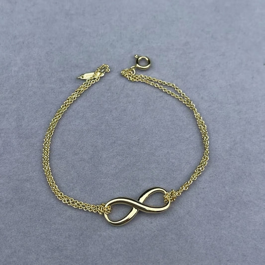 A delicate gold chain bracelet featuring a polished infinity symbol at the center, displayed on a textured gray background.