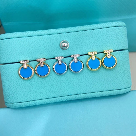 Six pairs of drop earrings with bright blue circular stones in gold, silver, and white gold settings, displayed on a textured turquoise jewelry box.