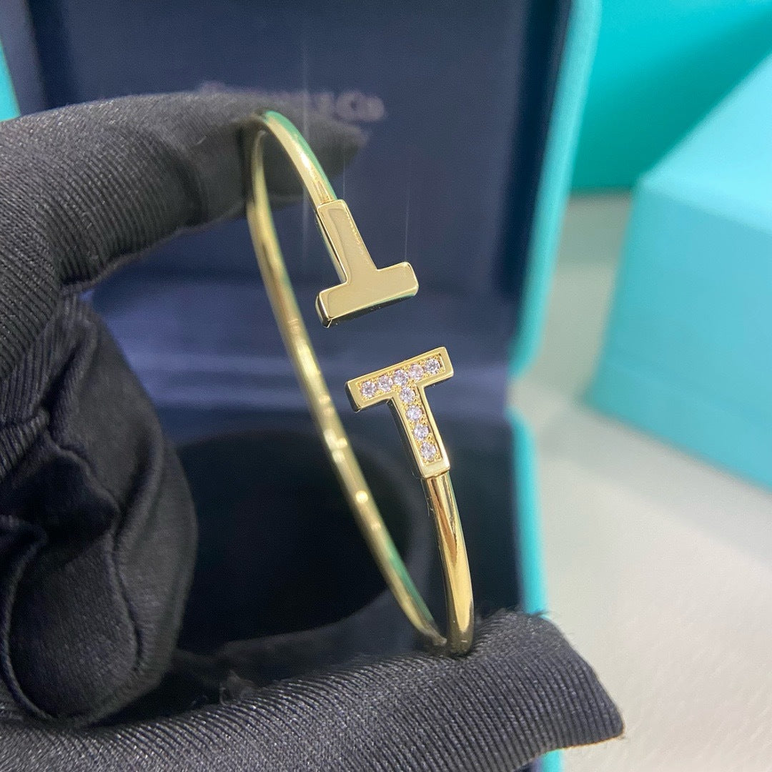 Gold bracelet with letter 'T' design held by a black glove against a blue background