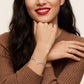 A close-up lifestyle shot of a woman wearing a delicate silver bracelet with three interlocking flower charms, one of which is light blue. She is wearing a brown ribbed sweater and has red lipstick on, with her other hand resting on her knee showing a matching silver ring.