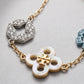 A close-up of a gold-tone chain bracelet featuring a mother-of-pearl four-leaf clover charm with a gold-tone monogram, a silver-tone pavé hexagon charm, and a partial light blue charm on a textured light gray background.