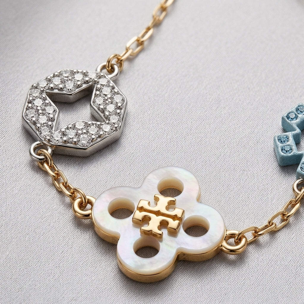 A close-up of a gold-tone chain bracelet featuring a mother-of-pearl four-leaf clover charm with a gold-tone monogram, a silver-tone pavé hexagon charm, and a partial light blue charm on a textured light gray background.