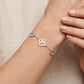 A delicate gold bracelet with three floral charms (one white, one light blue, and one clear crystal) worn on a woman's wrist against a neutral beige background.