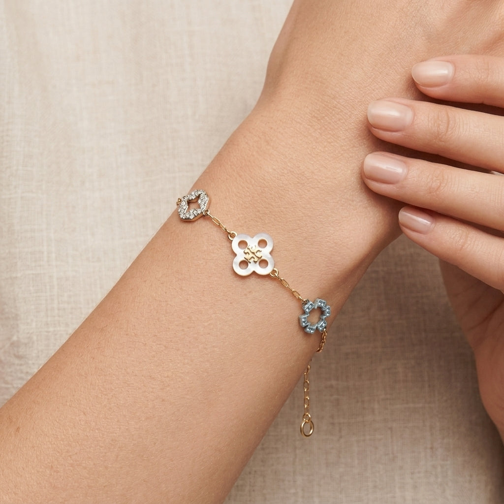 A delicate gold bracelet with three floral charms (one white, one light blue, and one clear crystal) worn on a woman's wrist against a neutral beige background.