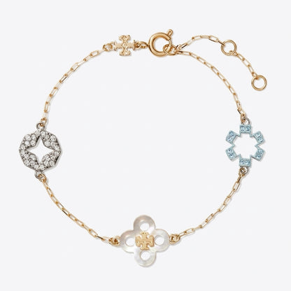 A delicate gold chain bracelet featuring three distinct flower-shaped charms: a central gold clover with a small gold cross in the center, a left-side silver star-shaped charm paved with clear stones, and a right-side light blue star-shaped charm paved with blue stones. The bracelet is displayed on a clean, light gray background.