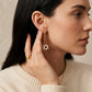 A close-up of a woman wearing a delicate gold dangle earring with a flower-shaped charm and a white pearl flower accent, paired with a matching gold ring and necklace.
