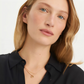 Close-up of a woman with blonde hair wearing a delicate gold necklace with a small flower charm, paired with a black collared shirt against a clean white background.
