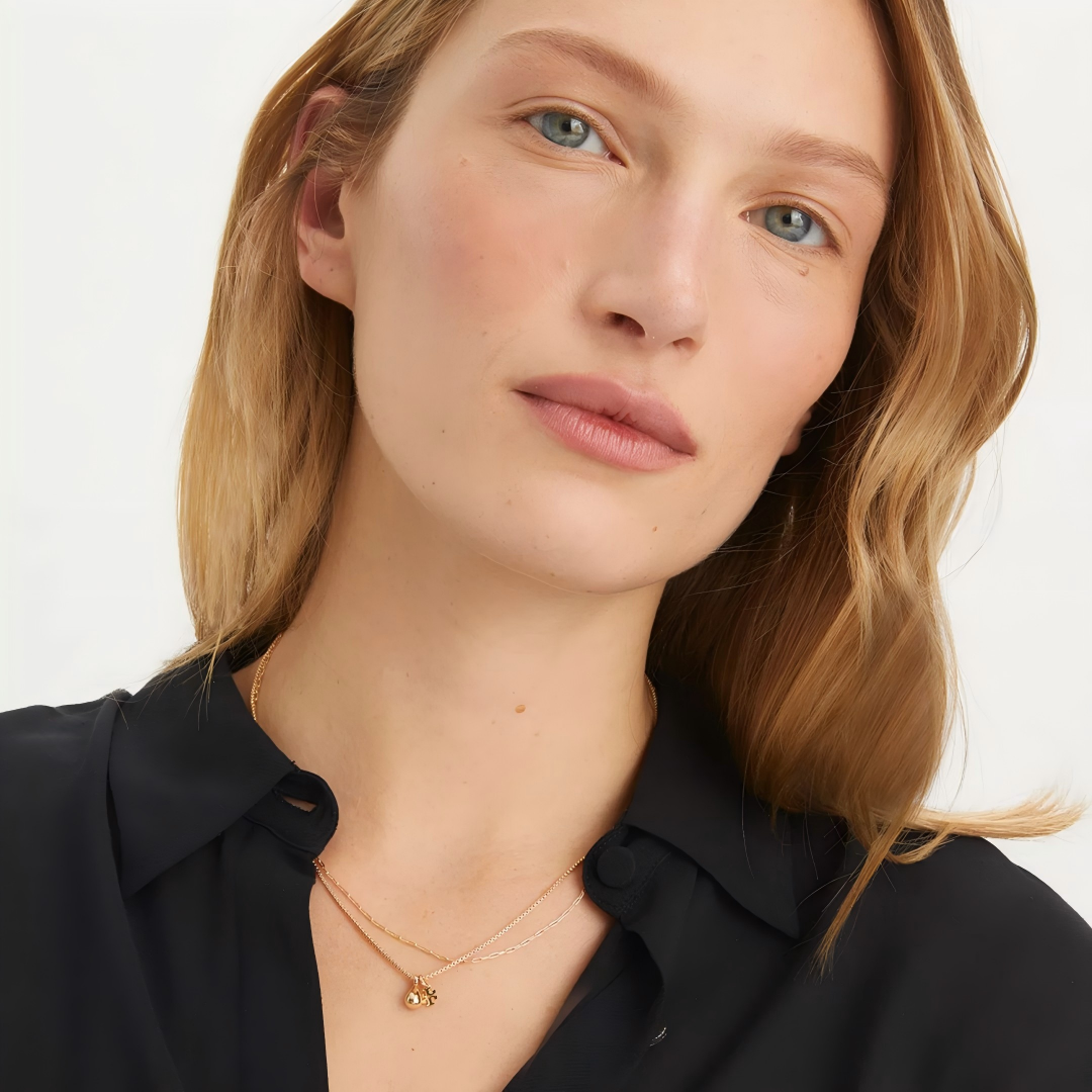 Close-up of a woman with blonde hair wearing a delicate gold necklace with a small flower charm, paired with a black collared shirt against a clean white background.