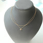 A delicate double-layered gold necklace with a small heart and flower pendant, displayed on a matte black jewelry bust.