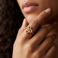 A close-up lifestyle shot of a model wearing a gold four-petal flower ring adorned with small clear crystals, set against a dark, softly blurred background.