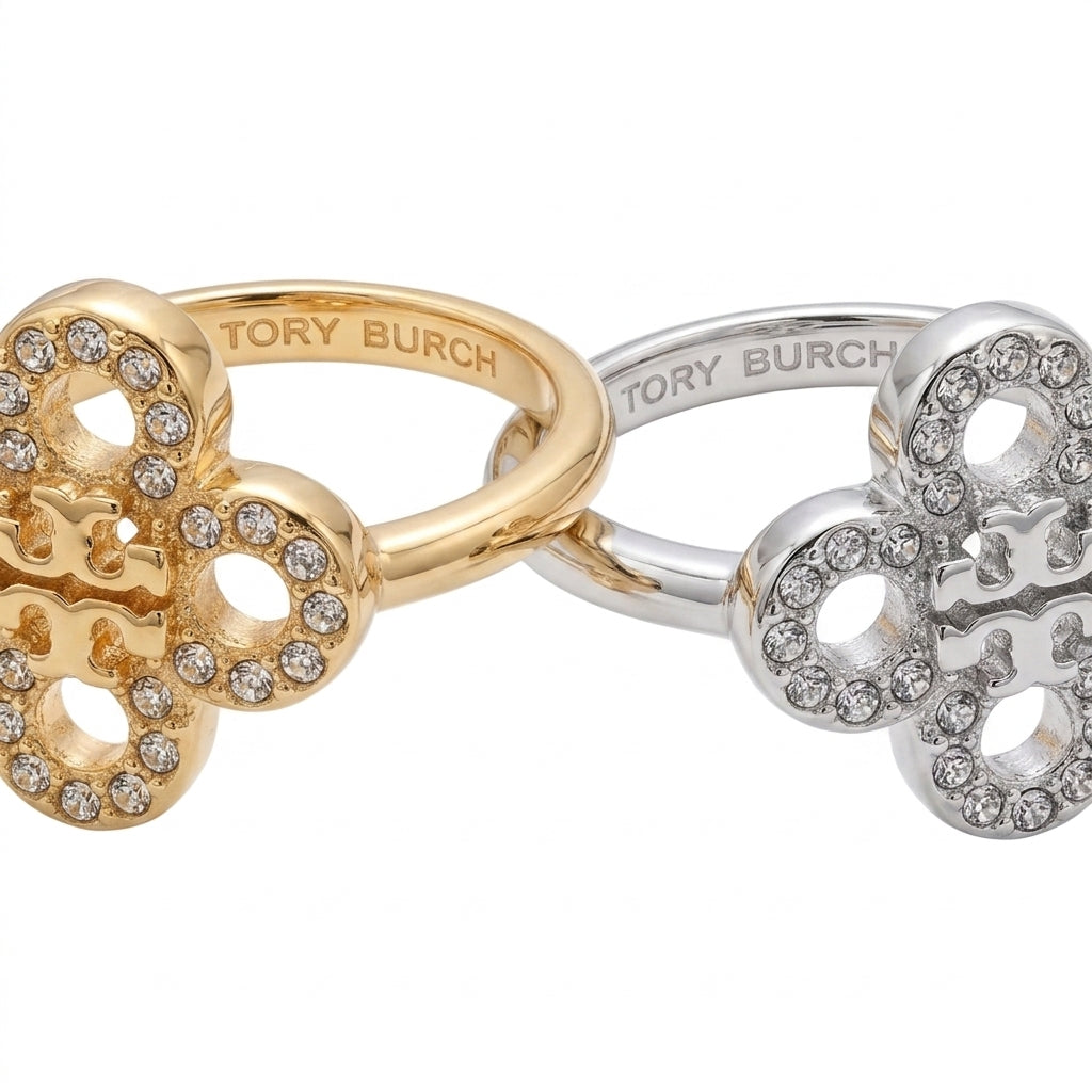 Two Tory Burch clover-shaped rings with pavé crystal detailing, one in yellow gold and one in silver, displayed on a clean white background.