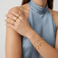 A close-up of a model wearing gold jewelry, including two stacked gold rings on one finger and three delicate gold chain bracelets with small heart and flower charms on the wrist, paired with a high-neck dusty blue satin top.